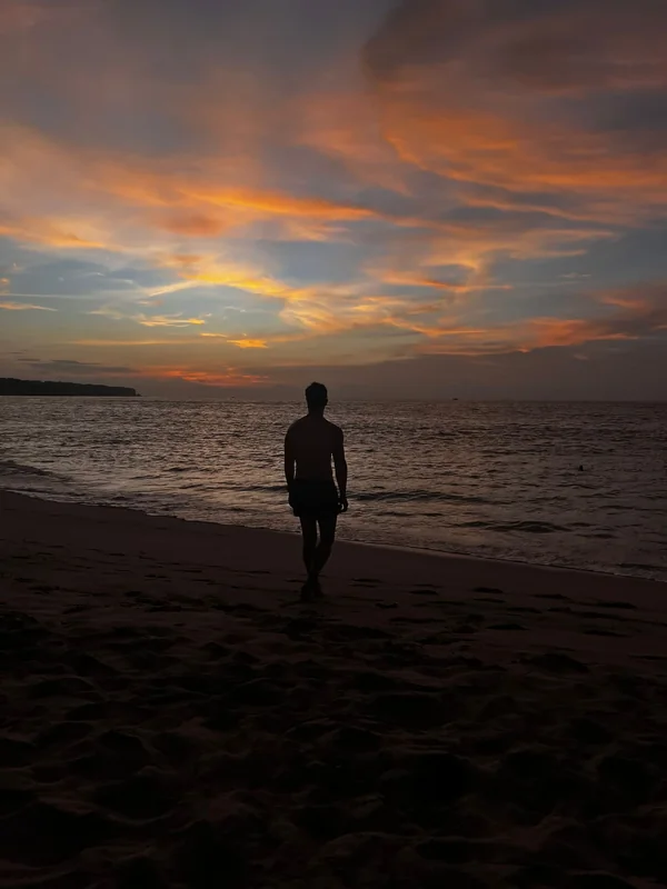 Silueta caminando descalzo por la arena de Jimbaran Beach durante un atardecer de cielo naranja en el sur de Bali