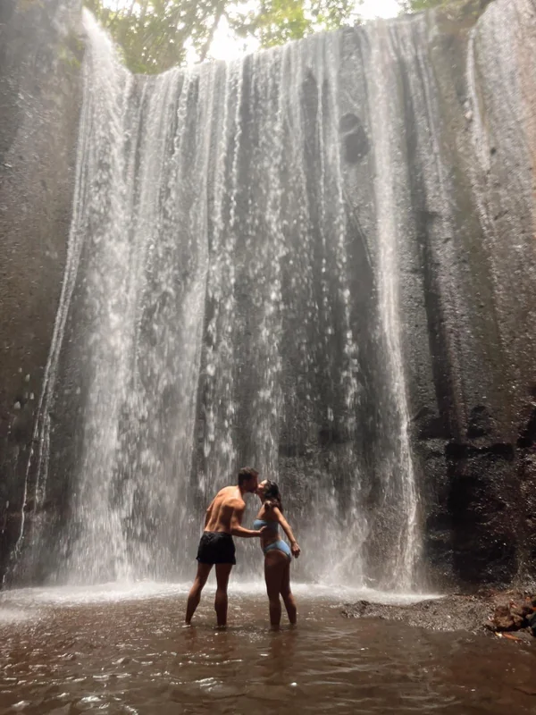 Pareja abrazada bajo la cortina de agua de la cascada Tukad Cepung, escondida en una gruta de roca en Bangli