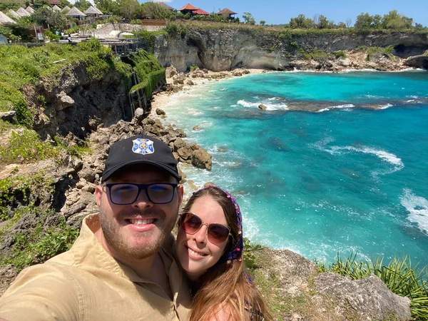 Pareja en selfie con los acantilados volcánicos y la cala turquesa de Devil's Tear en Nusa Lembongan al fondo