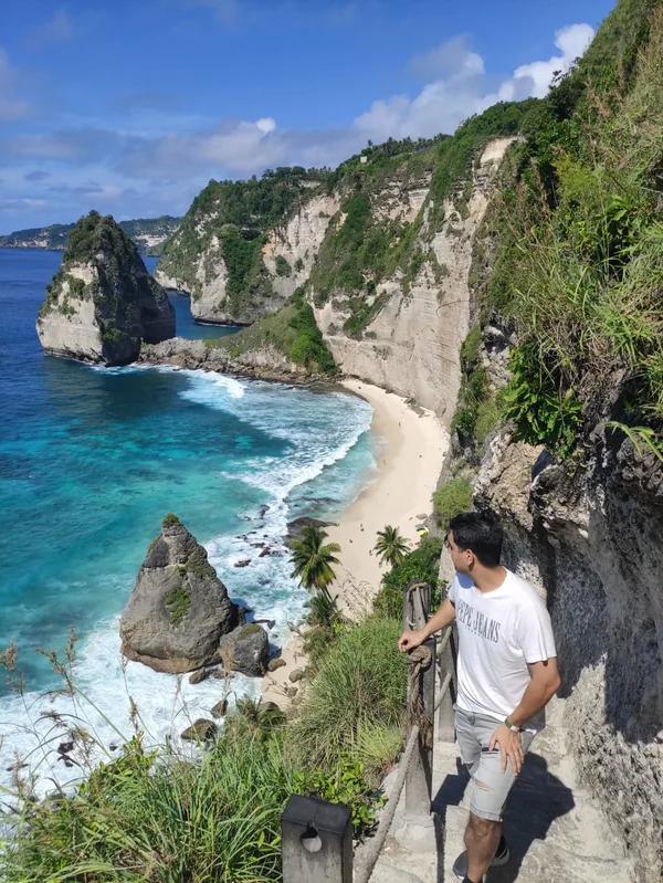 Hombre bajando por la escalera tallada en la roca con vista a la playa blanca y acantilados imponentes de Diamond Beach en Nusa Penida