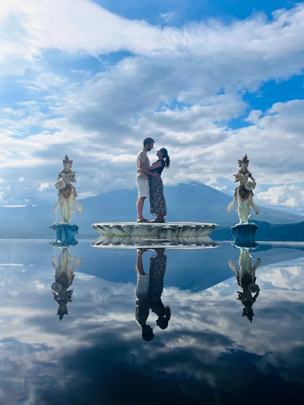 Pareja abrazada en la plataforma espejo con reflejo perfecto del volcán Monte Agung y estatuas balinesas a los lados