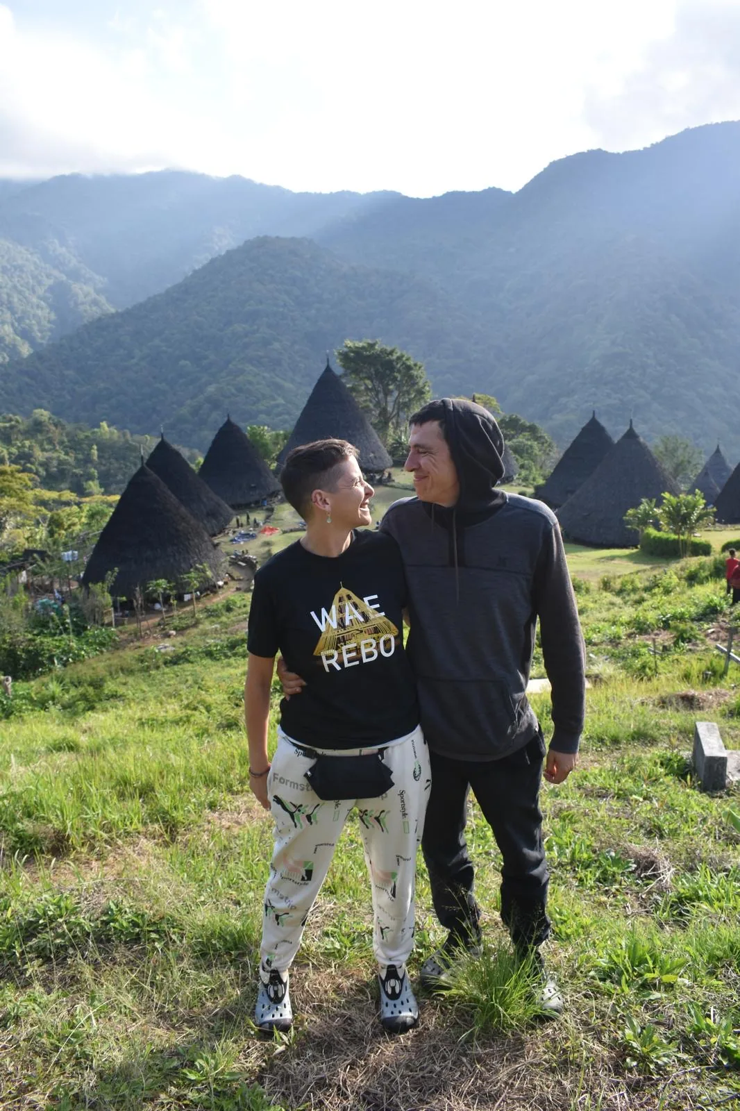 Pareja de viajeros de En Bali en la aldea de Wae Rebo con las casas mbaru niang al fondo